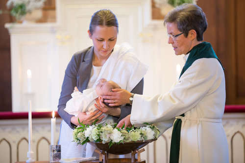 En mamma håller sitt barn över en dopfunt. En präst döper. Dopfunten är prydd med vita blommor